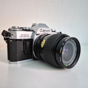 Könnte beinhalten: Eine silber-schwarze Canon AE-1 35mm Filmkamera mit einem schwarzen Objektiv, das die Aufschrift Super-Danubia MC 1:2.8 f=28mm und ein 52mm Filtergewinde trägt. Das Kameragehäuse hat einen strukturierten schwarzen Griff und eine silberne Deckkappe.