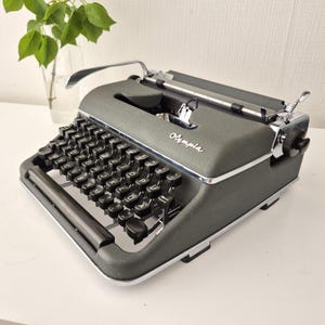 Könnte beinhalten: Vintage Olympia Schreibmaschine in dunkelgrauer Ausführung mit Chromverzierungen. Die Tastatur hat schwarze Tasten mit weißer Beschriftung. Das Wort "Olympia" ist vorne zu sehen. Ein klassisches Schreibwerkzeug.