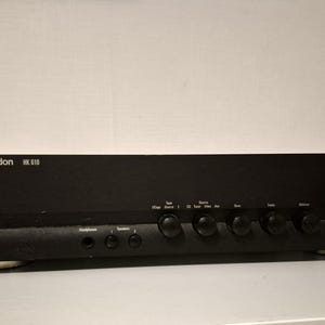 Op de afbeelding: Een zwarte Harman Kardon HK 610 geïntegreerde versterker met een aan-uitknop met een groen indicatielampje, een grote volumeknop en diverse kleinere draaiknoppen voor bronselectie, bas, hoge tonen en balans, evenals poorten voor hoofdtelefoons en luidsprekers.