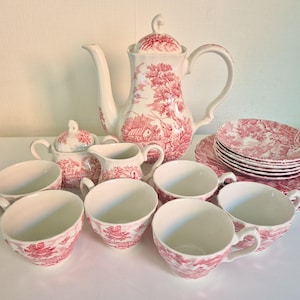 Op de afbeelding: Een theeservies bestaande uit een witte theepot, een suikerpot met deksel, een melkkan, zes theekopjes en zes schoteltjes. Elk onderdeel is versierd met een gedetailleerd rood toile-patroon met landelijke landschappen en gebouwen.