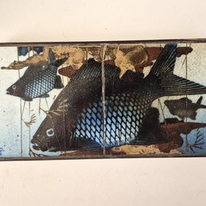 Könnte beinhalten: Rechteckiges Keramikfliesen-Set mit detaillierten Fischillustrationen. Die Fische sind überwiegend schwarz mit blauen Schuppenmustern, vor einem Hintergrund aus Hellblau und Braun. Die Fliesen sind von einem dunklen Metallrahmen eingefasst.