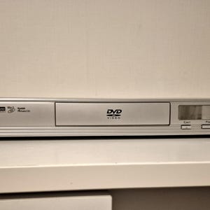 May include: A silver Motek DVD player with a horizontal rectangular design. The front panel features a power button with a green indicator light, a central disc tray labelled DVD Video, and a digital display currently showing the text disc.
