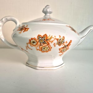 Può includere: Una teiera in porcellana bianca con un motivo floreale decorativo arancione e giallo con accenti neri. La teiera presenta un manico smerlato, un coperchio abbinato con pomello e un'ampia base esagonale con un sottile bordo dorato.