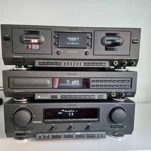 Op de afbeelding: Een stapel van drie zwarte Philips audiocomponenten. De bovenste unit is een dubbel cassettedeck, de middelste is een cd-speler en de onderste is een geïntegreerde versterker. Elk apparaat is voorzien van digitale displays, knoppen en draaiknoppen.