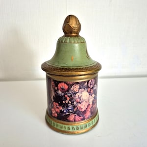 May include: A cylindrical lidded jar featuring a dark purple base decorated with a vibrant floral print of pink, red, and white flowers. The lid is light green with a gold-toned finial and gold-toned trim at the base and rim.