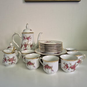 Könnte beinhalten: Ein Vintage-Teeservice mit Teekanne, Tassen, Untertassen, Sahnekännchen und Zuckerdose. Jedes Stück aus weißem Porzellan ist mit rosa Rosen und goldenen Verzierungen versehen. Das Set steht auf einer weißen Oberfläche.