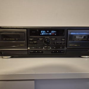 Puede incluir: Una pletina de casete estéreo Technics negra con dos compartimentos para cintas etiquetados como Deck 1 y Deck 2, ambos con función de auto-reverse. El panel central incluye una pantalla digital, un mando de volumen y varios botones.