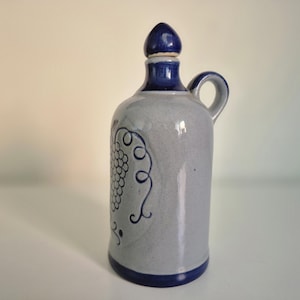 Può includere: Una brocca in ceramica grigio chiaro con manico, base e collo blu scuro. La brocca presenta un disegno in rilievo di un grappolo d'uva blu scuro sul davanti e include un tappo a punta blu scuro abbinato per l'apertura del collo.
