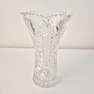Könnte beinhalten: Eine transparente, geschliffene Glasvase mit ausgestelltem Oberteil und komplizierten geometrischen Mustern. Die Vase hat ein Sternmuster auf dem Boden und detaillierte Schnitte am ganzen Körper, wodurch ein funkelnder Effekt entsteht. Die Vase ist etwa 20 cm hoch.