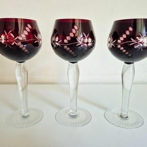 Può includere: Tre calici da vino con coppe color rosso mirtillo profondo caratterizzate da motivi floreali e geometrici intagliati. Ogni calice presenta uno stelo trasparente e liscio e una base circolare con un design a coste radiali sul bordo.