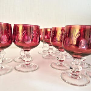 Pode incluir: Uma coleção de taças de vinho tinto rubi com hastes e bases transparentes e ornamentadas. As taças apresentam um design decorativo gravado e um interior dourado. As taças estão dispostas em fila sobre uma superfície branca.