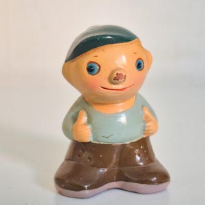Pode incluir: Uma pequena estatueta de um personagem com rosto cor de pêssego, olhos azuis e um pequeno nariz vermelho. A figura usa um boné verde-azulado escuro, uma camisa azul clara e sapatos castanhos. O personagem tem uma expressão sorridente.