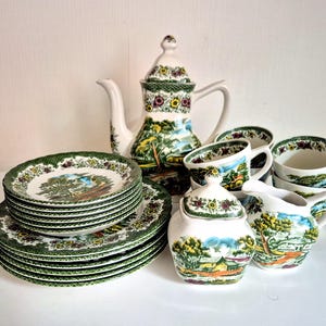 Op de afbeelding: Een keramische theeset met een witte basis en kleurrijke landschapsillustraties van bomen, gebouwen en heuvels. De set bevat een hoge theepot met deksel, een suikerpot met deksel, een melkkannetje, diverse theekopjes en een stapel bijpassende borden.