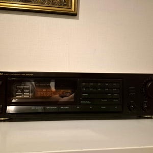 Op de afbeelding: Een zwarte Onkyo Quartz Synthesized Tuner-versterker met een centraal digitaal display, meerdere rechthoekige drukknoppen, een grote ronde volumeknop aan de rechterkant en een hoofdtelefoonaansluiting aan de linkerkant van het voorpaneel.