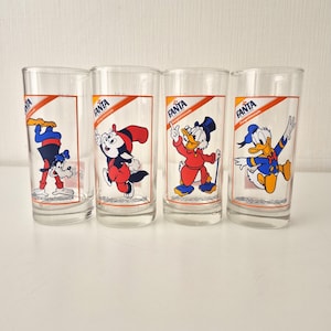Könnte beinhalten: Set aus vier Trinkgläsern mit Disney-Zeichentrickfiguren. Jedes Glas hat ein Fanta-Logo und eine andere Figur: Goofy, eine fuchsartige Figur, Dagobert Duck und Donald Duck. Die Gläser stehen auf einer weißen Oberfläche.