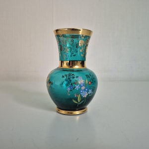 Pode incluir: Um vaso de vidro verde-azulado com corpo bulboso e gargalo alargado, decorado com bordas douradas na abertura, no gargalo e na base. A superfície é adornada com motivos florais pintados em azul, branco e verde, além de folhas douradas.