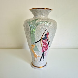 Puede incluir: Un jarrón de cerámica blanca con superficie texturizada, borde y base con acabado dorado. El diseño muestra una figura de silueta negra con un vestido rosa estampado, un árbol verde y un pequeño animal de color beige en relieve.