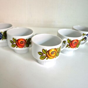 Op de afbeelding: Een set van vijf witte keramische kopjes, elk voorzien van een handgeschilderd bloemmotief met gestileerde rozen in blauwe, oranje en gele tinten, vergezeld van groene bladeren. De kopjes hebben een gladde binnenkant en een geribbelde voet.