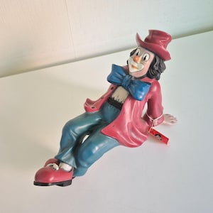 Könnte beinhalten: Eine Clown-Figur in liegender Position, bekleidet mit einem roten Zylinder, einer passenden roten Jacke, einer großen blauen Fliege und einer blaugrünen Hose. Der Clown hat ein weißes Gesicht mit bemalten Zügen, schwarzes Haar und rote Schuhe.