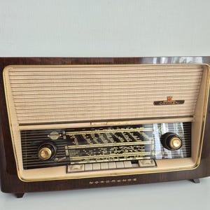 Könnte beinhalten: Ein Vintage-Radio vom Typ Nordmende Carmen mit einem Gehäuse aus dunklem Holzfurnier, einem cremefarbenen Stofflautsprechergitter und einer schwarzen Glasskala mit goldfarbenen Drehknöpfen. Der Markenname und das Modell sind in Gold aufgedruckt.