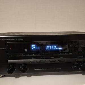 Può includere: Un ricevitore stereo audio-video Kenwood nero modello KR-V8050 dotato di un display digitale centrale che mostra 87.50, una grande manopola del volume sulla destra, vari pulsanti funzione, selettori di ingresso e un jack per cuffie sul pannello.