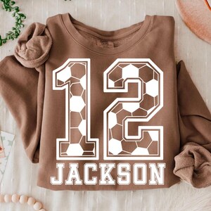 K&ouml;nnte beinhalten: Ein braunes Langarm-Sweatshirt mit einer gro&szlig;en wei&szlig;en Grafik der Zahl 12, die mit einem Fu&szlig;ballmuster gef&uuml;llt ist. Unter der Zahl 12 ist der Name JACKSON in einer fetten wei&szlig;en Serifenschrift auf der Brustpartie aufgedruckt.