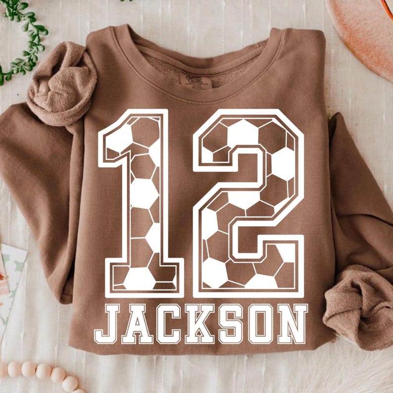 K&ouml;nnte beinhalten: Ein braunes Langarm-Sweatshirt mit einer gro&szlig;en wei&szlig;en Grafik der Zahl 12, die mit einem Fu&szlig;ballmuster gef&uuml;llt ist. Unter der Zahl 12 ist der Name JACKSON in einer fetten wei&szlig;en Serifenschrift auf der Brustpartie aufgedruckt.