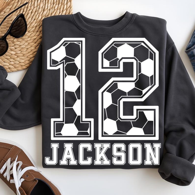 K&ouml;nnte beinhalten: Ein dunkelgraues Langarm-Sweatshirt mit einer gro&szlig;en Nummer 12, die in einem schwarz-wei&szlig;en Fu&szlig;ballmuster bedruckt ist. Unter der Nummer ist der Name JACKSON in fetten, wei&szlig;en Gro&szlig;buchstaben mit schwarzer Umrandung auf der Brust aufgedruckt.