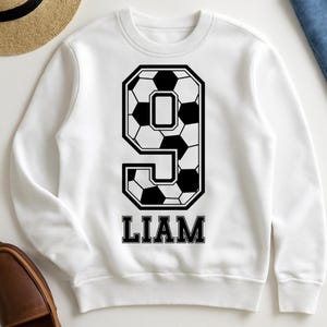 K&ouml;nnte beinhalten: Ein wei&szlig;es Langarm-Sweatshirt mit einer gro&szlig;en schwarz-wei&szlig;en Grafik der Zahl Neun, die mit einem Fu&szlig;ballmuster gef&uuml;llt ist. Unter der Zahl ist der Name LIAM in einer fetten, schwarzen Serifenschrift mittig auf der Brust aufgedruckt.