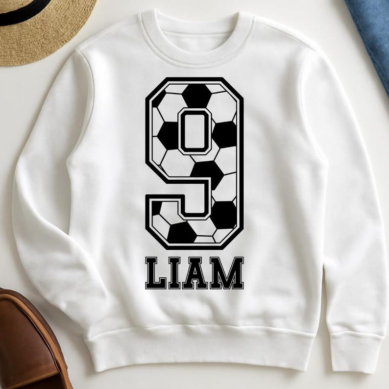 K&ouml;nnte beinhalten: Ein wei&szlig;es Langarm-Sweatshirt mit einer gro&szlig;en schwarz-wei&szlig;en Grafik der Zahl Neun, die mit einem Fu&szlig;ballmuster gef&uuml;llt ist. Unter der Zahl ist der Name LIAM in einer fetten, schwarzen Serifenschrift mittig auf der Brust aufgedruckt.