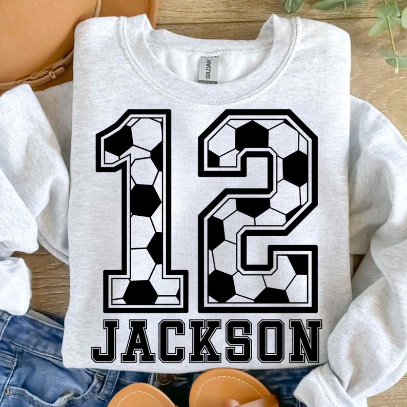 K&ouml;nnte beinhalten: Ein hellgraues Sweatshirt mit Rundhalsausschnitt und der gro&szlig;en Zahl 12, die mit einem schwarz-wei&szlig;en Fu&szlig;ballmuster bedruckt ist. Unter der Zahl steht der Name JACKSON in fetten, schwarzen Gro&szlig;buchstaben. Das Kleidungsst&uuml;ck hat lange &Auml;rmel.