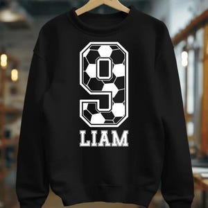 K&ouml;nnte beinhalten: Ein schwarzes Langarm-Sweatshirt mit einer gro&szlig;en wei&szlig;en Grafik der Zahl 9, die mit einem schwarz-wei&szlig;en Fu&szlig;ballmuster gef&uuml;llt ist. Unter der Zahl ist der Name LIAM in einer fetten, wei&szlig;en Schriftart in Gro&szlig;buchstaben aufgedruckt.