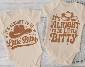 Könnte beinhalten: Zwei beige kurzärmelige Babybodys mit braunen Grafikdrucken von Cowboyhüten und dem Text It's Alright To Be Little Bitty. Die Designs enthalten kleine Sternakzente und eine geschwungene Typografie auf hellem Stoff.