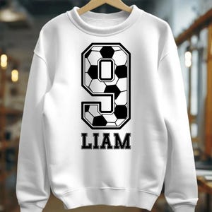K&ouml;nnte beinhalten: Ein wei&szlig;es Langarm-Sweatshirt mit Rundhalsausschnitt und einer gro&szlig;en Grafik der Zahl Neun, die mit einem schwarz-wei&szlig;en Fu&szlig;ballmuster gef&uuml;llt ist. Unter der Zahl Neun ist der Name LIAM in einer fetten, schwarzen Serifenschrift gedruckt.
