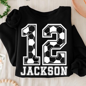 K&ouml;nnte beinhalten: Ein schwarzes Langarm-Sweatshirt mit einer gro&szlig;en Grafik der Zahl 12, die mit einem schwarz-wei&szlig;en Fu&szlig;ballmuster gef&uuml;llt ist. Unter der Zahl ist der Name JACKSON in fetten, wei&szlig;en Gro&szlig;buchstaben mit schwarzer Umrandung aufgedruckt.