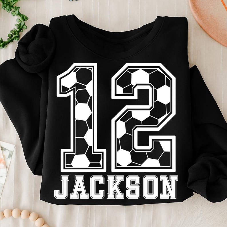 K&ouml;nnte beinhalten: Ein schwarzes Langarm-Sweatshirt mit einer gro&szlig;en Grafik der Zahl 12, die mit einem schwarz-wei&szlig;en Fu&szlig;ballmuster gef&uuml;llt ist. Unter der Zahl ist der Name JACKSON in fetten, wei&szlig;en Gro&szlig;buchstaben mit schwarzer Umrandung aufgedruckt.