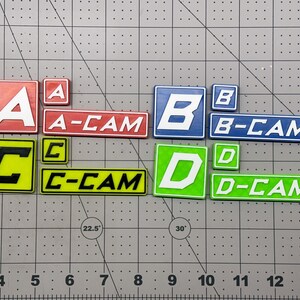Op de afbeelding: Een verzameling rechthoekige en vierkante plastic labels met de letters A, B, C en D in verschillende kleuren. Elke set bevat een grote letter, een kleine letter en een rechthoekig label met A-CAM, B-CAM, C-CAM of D-CAM.