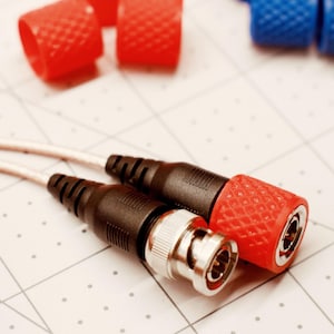 May include: Close-up of two black and silver BNC connectors with white cables, and red and blue protective caps. The red caps have a textured surface. The background is a white grid.
