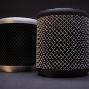 May include: Two cylindrical objects with a geometric lattice design. One is black with a silver rim, the other is black with a dark gray lattice. The objects are on a dark gray surface.