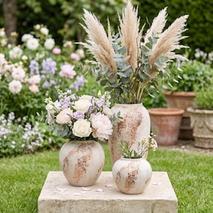 Peut inclure: Trois vases décoratifs de différentes tailles, avec une base crème et un motif tourbillonnant cuivré. Ils contiennent des compositions florales variées : herbe de la pampa, eucalyptus et fleurs blanches et lavande. La scène se déroule en extérieur.