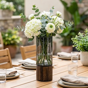 Peut inclure: Un vase bicolore avec une base en bois foncé et une partie supérieure en verre transparent. Le vase contient un bouquet de fleurs blanches et de feuillage vert. Le vase est sur une table en bois, avec des assiettes et des verres à proximité.
