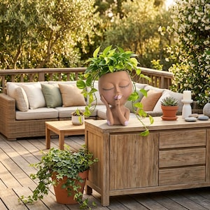 Peut inclure: Un pot de fleurs en céramique brun clair en forme de tête humaine avec les yeux fermés et un doigt posé sur les lèvres dans un geste de silence. Le pot contient une plante pothos verte retombante et repose sur un meuble en bois.