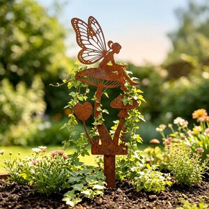 Peut inclure: Une décoration de jardin en métal couleur rouille représentant la silhouette d'une fée aux ailes détaillées assise sur un grand champignon, accompagnée d'un plus petit champignon et d'une feuille, fixée sur un piquet vertical à planter.