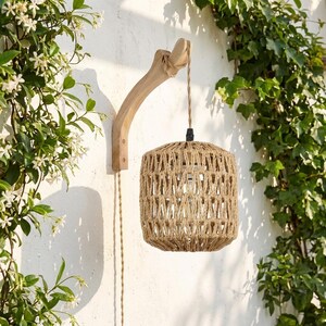 Peut inclure: Une applique murale composée d'un support en bois clair courbé et d'un abat-jour cylindrique en corde de jute naturelle tressée. L'abat-jour est suspendu par un cordon torsadé enroulé autour du bras en bois, créant un effet de suspension.
