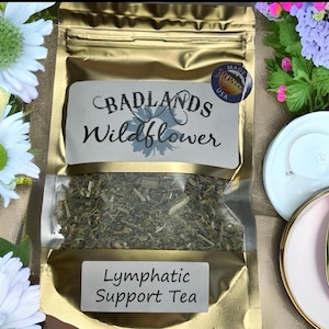 Op de afbeelding: Een goudkleurig zakje Badlands Wildflower Lymphatic Support Tea. Het zakje heeft een doorzichtig venster met gedroogde kruiden. Het etiket luidt "Badlands Wildflower" en "Lymphatic Support Tea". Een sticker "Made in Montana USA" is zichtbaar.