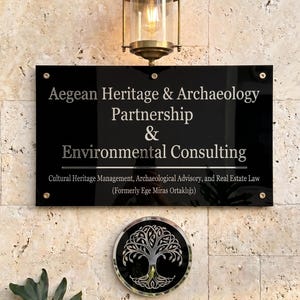 May include: A black sign with white text reading "Aegean Heritage & Archaeology Partnership & Environmental Consulting." Below the sign is a circular emblem with a tree design. A brass lantern hangs above the sign on a stone wall.