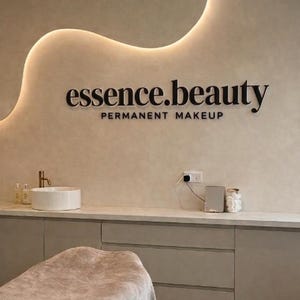 May include: The interior of a beauty salon features a wall with the words "essence.beauty" and "PERMANENT MAKEUP" in black lettering. A white sink, bottles, and a gold faucet are visible. A beige massage table is in the foreground.