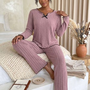 Può includere: Completo pigiama in maglia a coste malva. La parte superiore a maniche lunghe ha uno scollo smerlato con un piccolo fiocco nero e finiture nere. I pantaloni abbinati hanno una gamba larga e finiture nere. La modella è seduta su un letto.