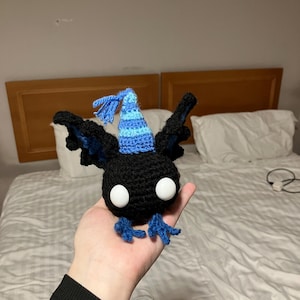 May include: A handmade black crochet bat with blue accents, large white eyes, and a blue and light blue party hat. The bat has outstretched wings and blue feet. The plush toy is held in a hand.