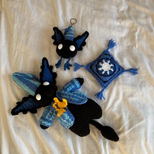May include: A collection of handmade crochet items, including a black and blue creature with white eyes, a small keychain creature, and a blue square with a white star design. The items are arranged on a white surface.